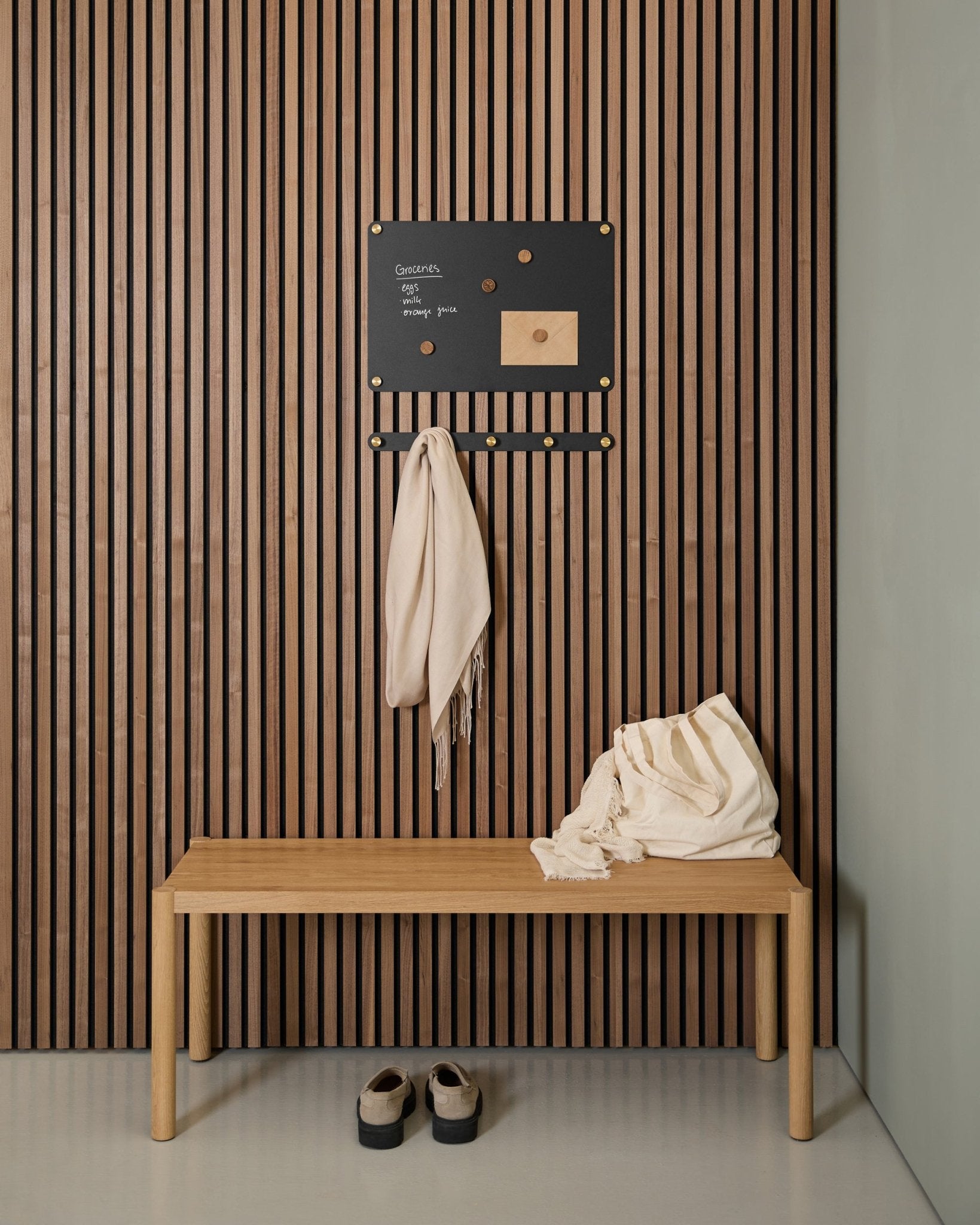 Minimalist entryway with a wooden bench, two scarves, sandals, and a small WoodUpp Norr Magnetic Board on an Akupanel acoustic wood slatwall panel.